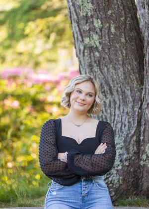 Young blonde woman in black sheer top and jeans poses confidently with arms crossed beside a large tree outdoors.