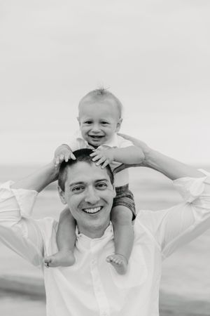 Black and white photo of a smiling father with a happy baby sitting on his shoulders at the beach.