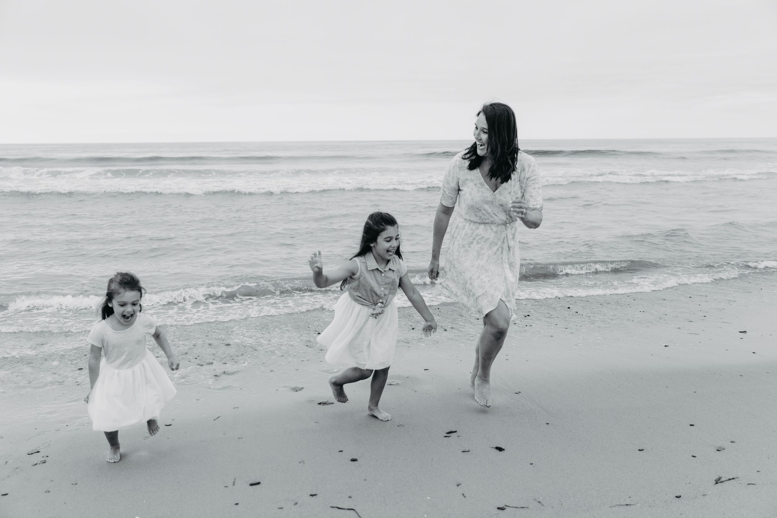 Black and white photo of mother and two daughters in white dresses running on beach.