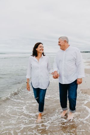 Happy mature couple holding hands walking barefoot in ocean waves on cloudy beach day.