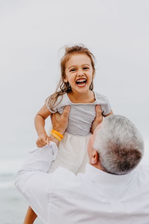Laughing little girl in gray dress is lifted joyfully by a man near the ocean beach.