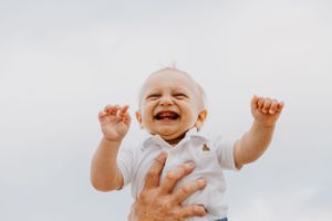 Joyful baby in white outfit laughs with arms raised against a bright sky background.