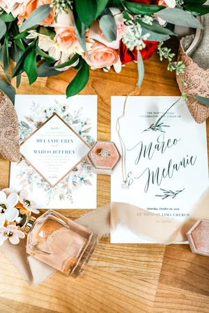 Elegant wedding invitation suite with floral details, ring box, perfume, and peach roses on a wooden table.
