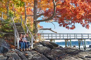 Couple sits on rocky shoreline beneath vibrant red autumn foliage beside a waterfront bridge.