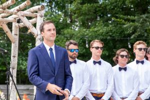 Groom in navy suit stands at outdoor wedding altar with groomsmen in white shirts and bow ties.