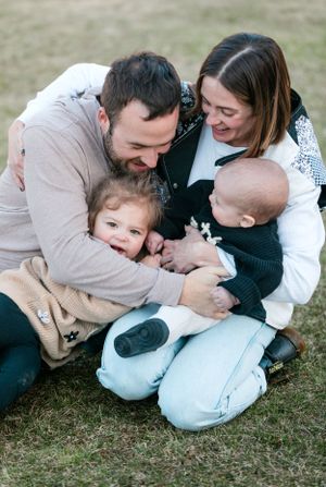 A loving family of four sitting on grass, parents cuddling toddler and baby outdoors.