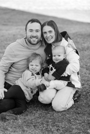 Black and white family portrait of parents smiling with two young children sitting on grass outdoors.