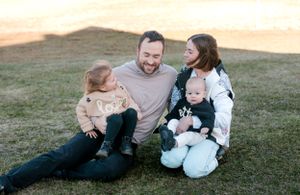 Happy family of four sitting on grass outdoors, parents smiling with toddler and baby in casual setting.