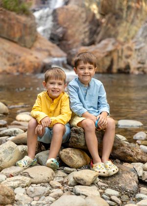 Two smiling boys in colorful shirts sitting on rocks in front of a waterfall.