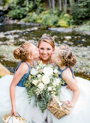 Smiling bride holding white bouquet being kissed on cheeks by two flower girls in blue dresses.