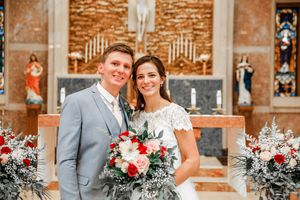 Bride and groom smiling together at church altar with red and white floral arrangements during wedding ceremony.