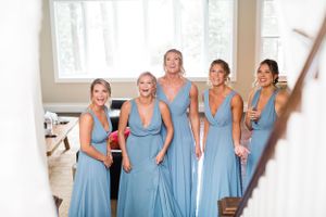 Five bridesmaids in dusty blue dresses smiling and reacting joyfully indoors at a wedding.