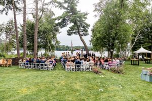 Outdoor lakeside wedding ceremony with guests seated on white chairs surrounded by tall pine trees.