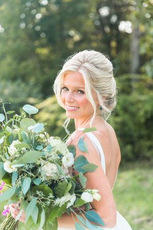 Smiling bride with blonde updo holding lush green and white floral bouquet outdoors in natural light.