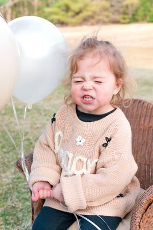Toddler girl in beige knit sweater making a funny face while holding white balloons outdoors.