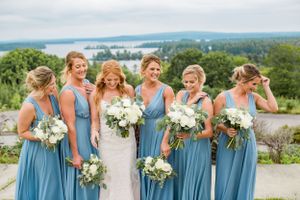 Bride with five bridesmaids in blue dresses holding white bouquets with lake view behind.