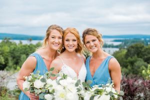 Bride with two bridesmaids in blue dresses holding white bouquets with scenic lake view behind them.