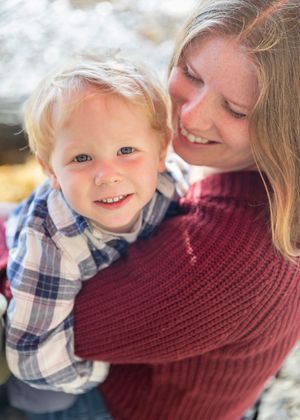 A smiling blonde toddler boy being held by his mother in a red knit sweater outdoors.