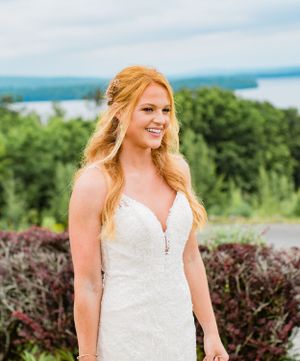 Radiant bride in lace wedding gown smiles outdoors with scenic lake and greenery behind her.