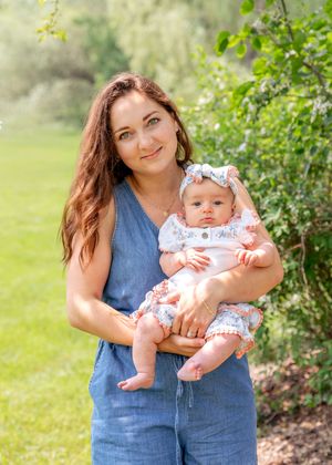 A young mother holding her newborn baby girl in a white floral outfit outdoors in a green garden.