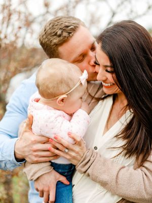 Loving parents kissing their baby girl on cheeks outdoors in soft natural light during family portrait.