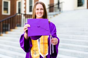 Smiling female graduate in purple cap and gown holding diploma on university steps.
