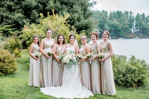Bride with six bridesmaids in champagne gowns holding white bouquets outdoors by a lake.