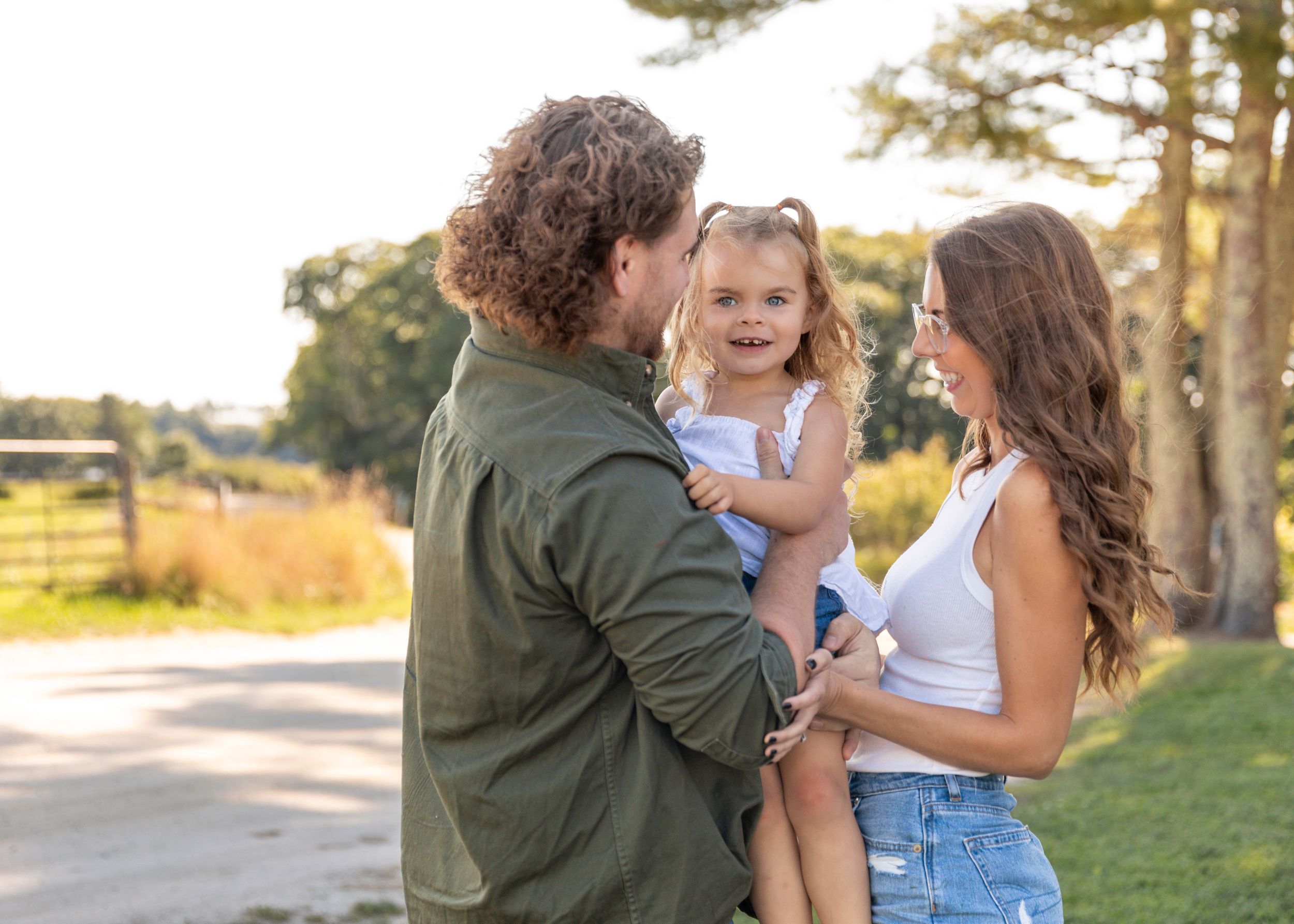 Young family with curly-haired dad holding smiling toddler girl with mom on a sunny rural farm path.