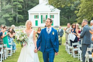 Newlywed couple walking down outdoor aisle holding hands after wedding ceremony near white chapel.