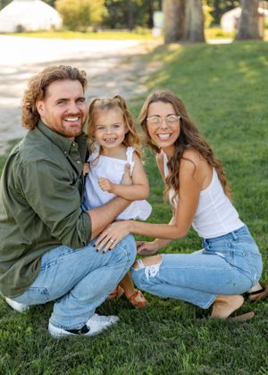 Happy family of three posing on green grass, parents smiling with young daughter in white dress.