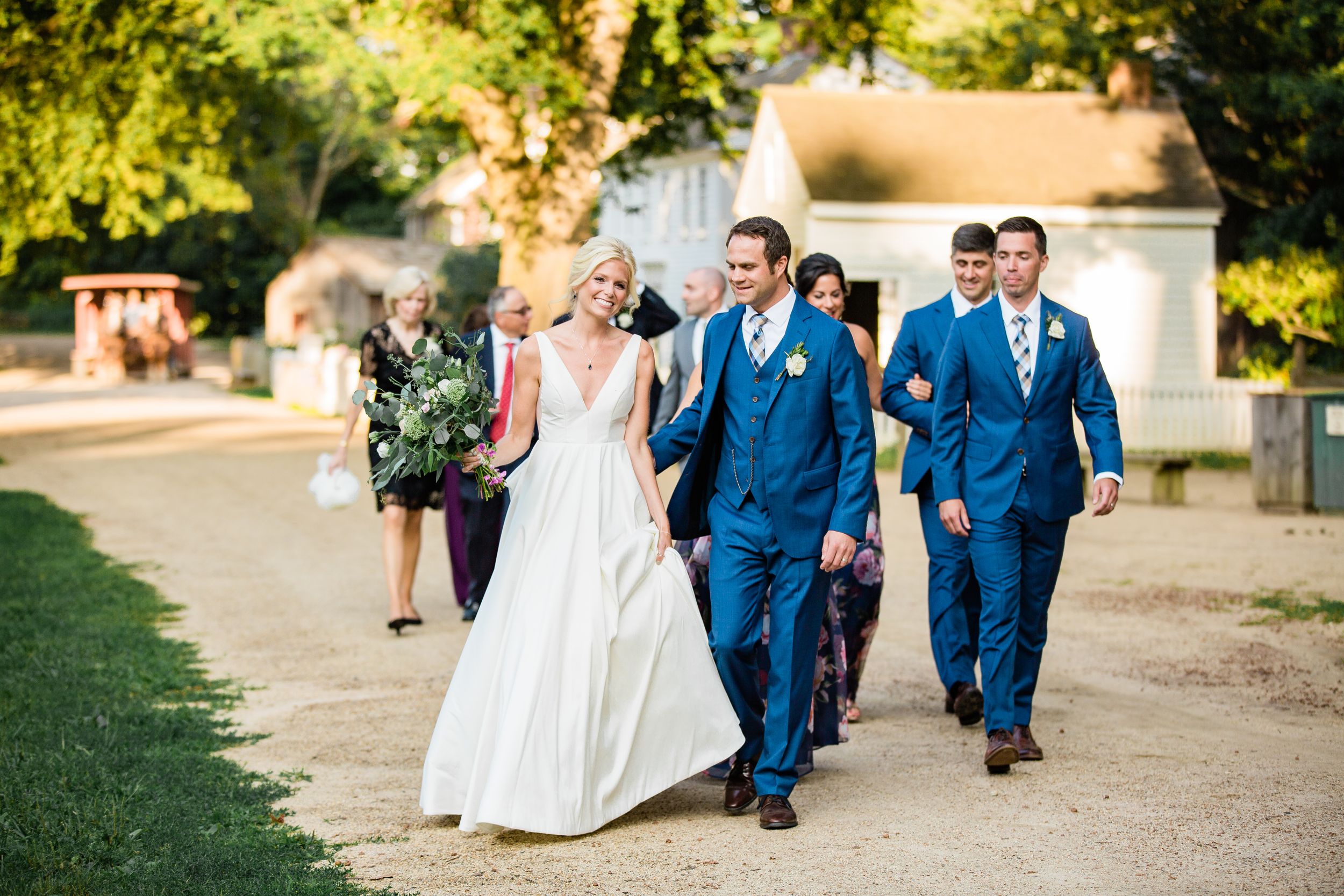 Wedding party walking outdoors in blue suits and white bridal gown on sunny gravel path.