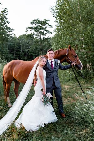 A bride and groom posing with a chestnut horse in a lush green field on their wedding day.
