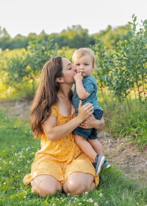 Mother in yellow dress kissing toddler son while sitting on green grass in sunny garden.