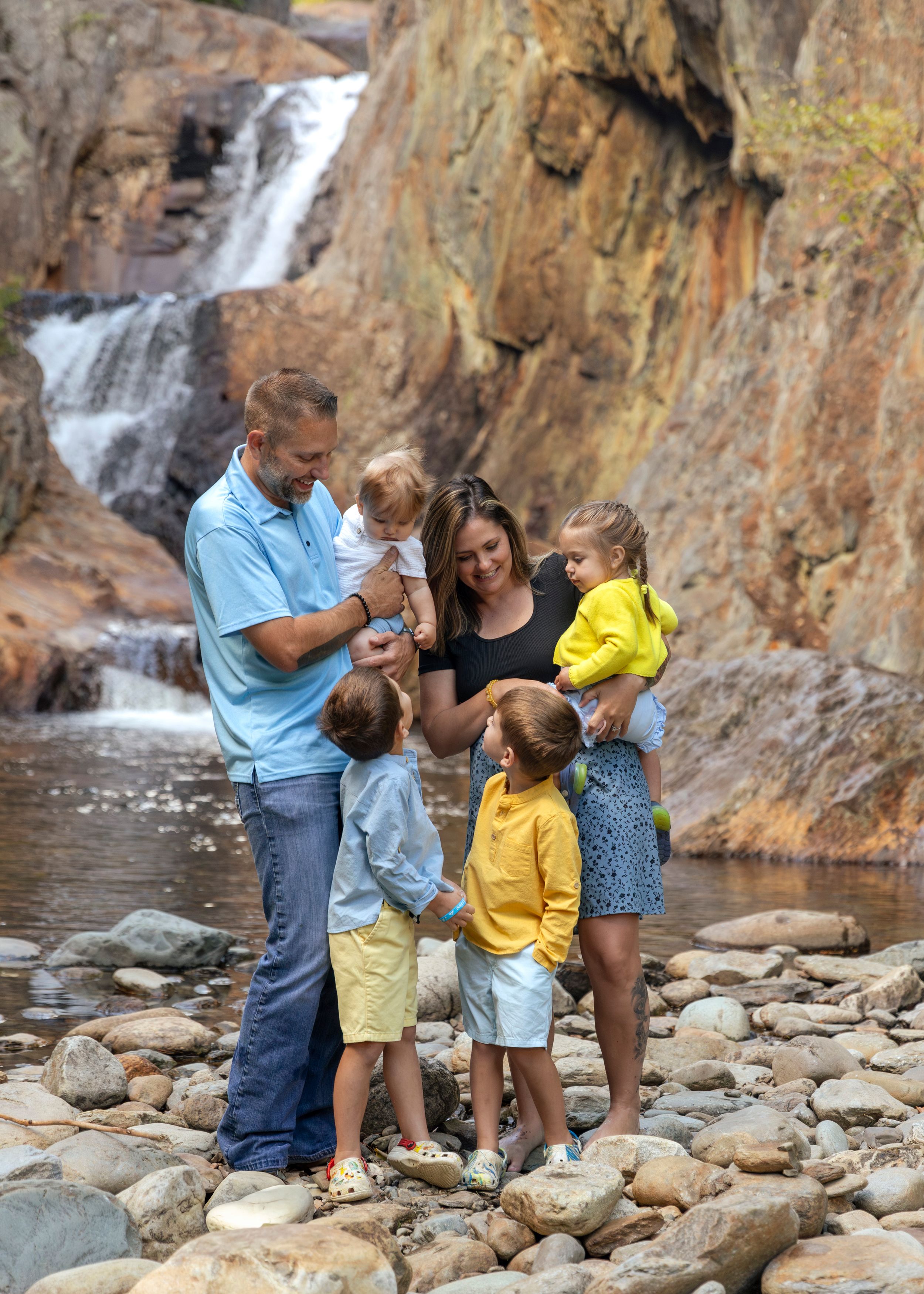 Family of six with four young children gathered by a scenic waterfall on rocky terrain outdoors.