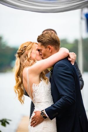Bride and groom sharing first kiss at waterfront wedding ceremony under a draped arch.