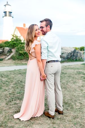Romantic couple holding hands near historic lighthouse, woman in blush pink flowing gown smiling.
