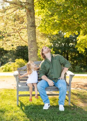 Father and toddler daughter sharing a playful moment on a park bench under green trees.