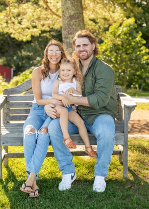 A happy young family of three sitting on a wooden bench in a sunny green park setting.