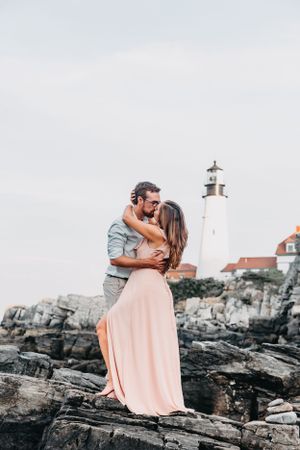 A couple sharing a romantic kiss on rocky coastal cliffs with a classic lighthouse in the background.