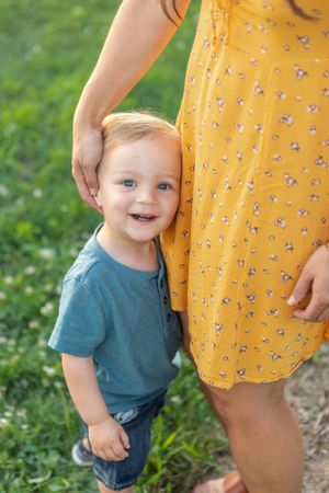 A smiling toddler boy in a blue shirt leaning against his mothers yellow floral dress outdoors.