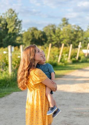 Young mother in yellow dress holding toddler son while gazing upward on a farm path.