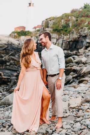 A romantic couple holding hands on a rocky beach with a lighthouse in the background.