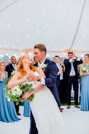 Groom dips smiling bride holding white bouquet during first dance under a tent with string lights.