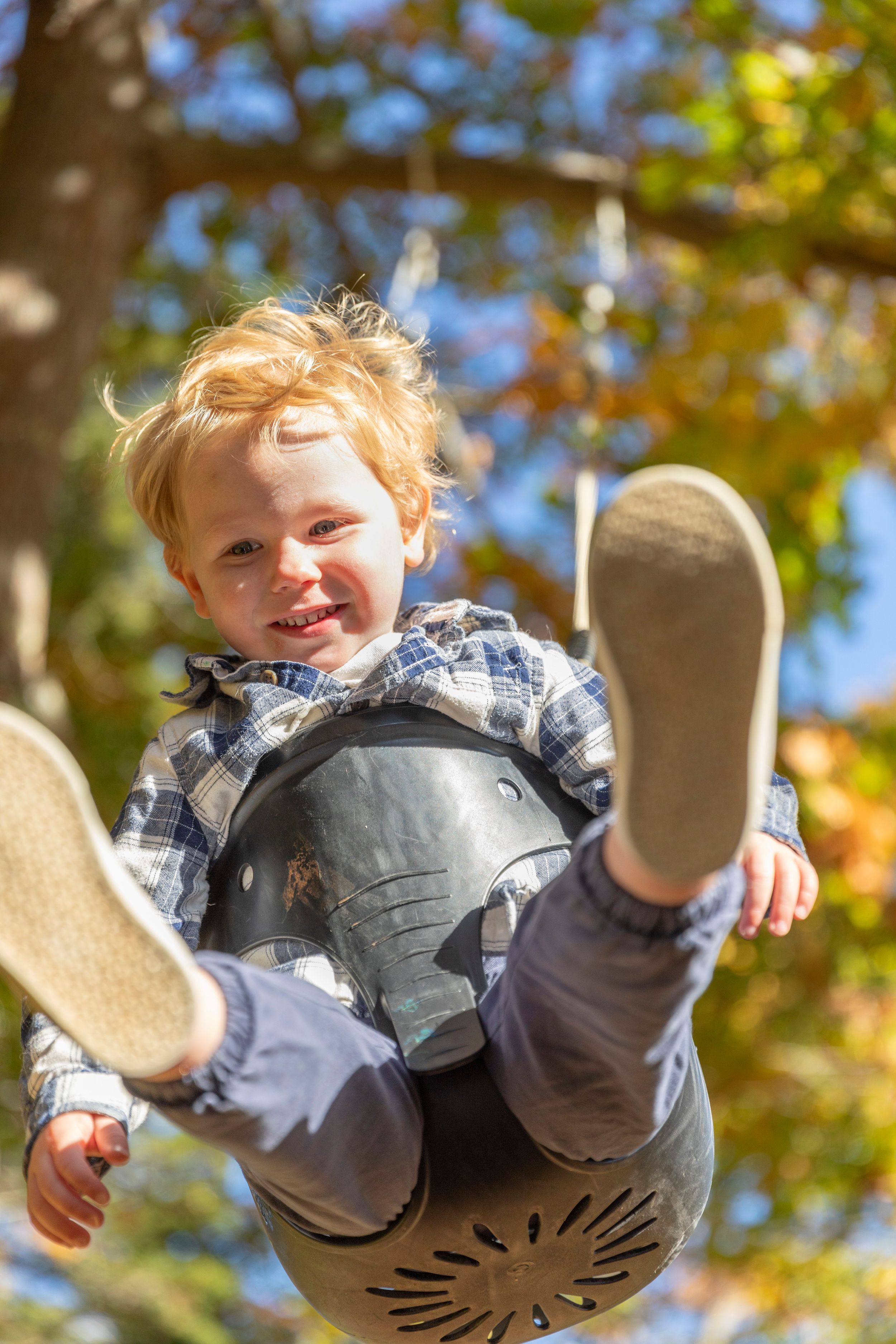 Smiling red-haired toddler boy swinging on a bucket swing surrounded by colorful autumn trees outdoors.