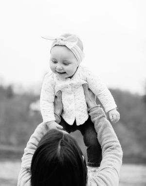 Black and white photo of laughing baby in bow headband being lifted up by parent outdoors.