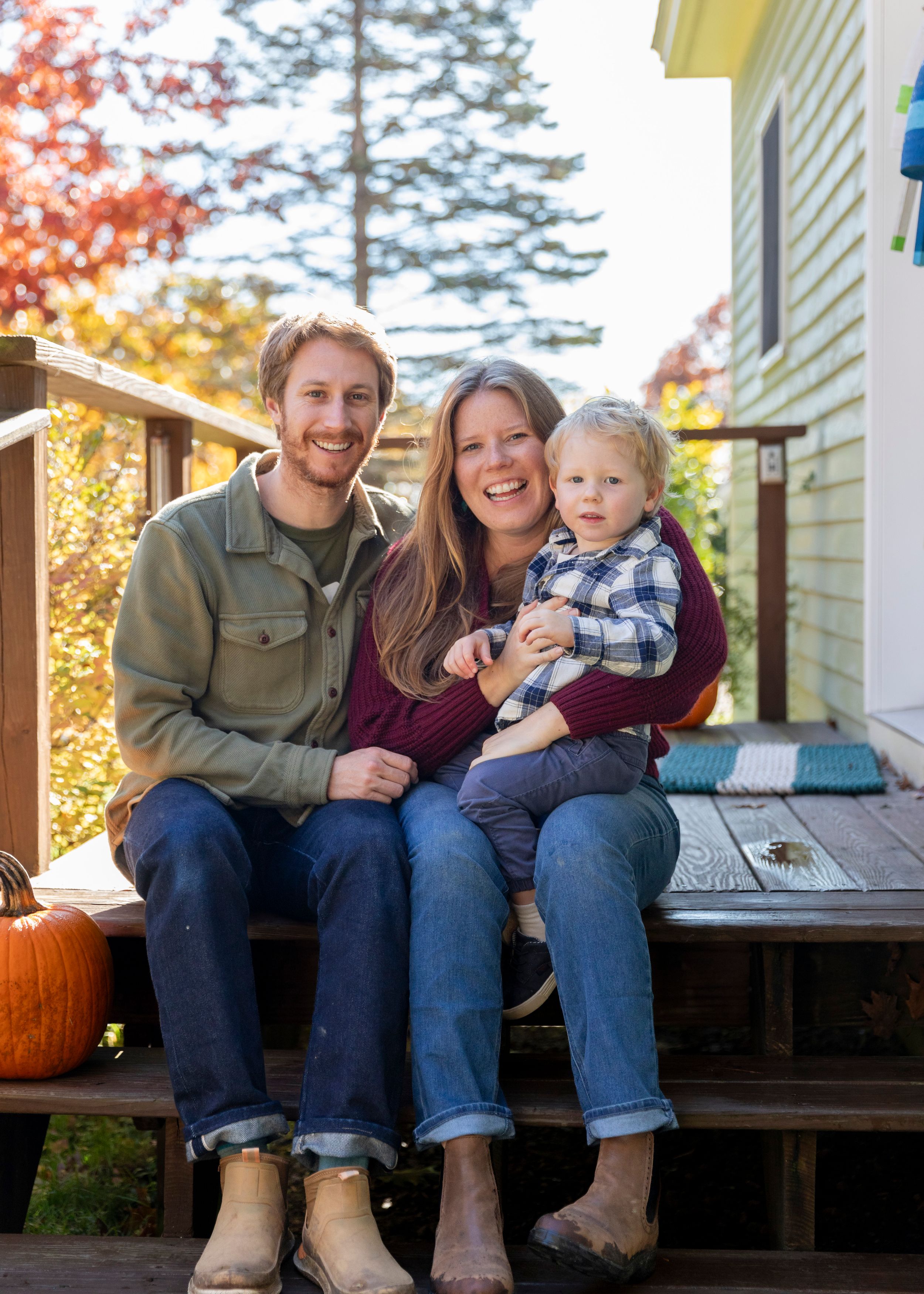 Young family with toddler sitting on porch steps surrounded by autumn foliage and pumpkins.