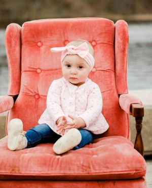 Adorable baby girl in pink outfit and bow headband sitting in a vintage coral velvet chair.