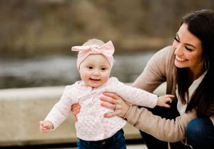 Smiling mother helps happy baby girl in pink floral outfit take first steps outdoors.