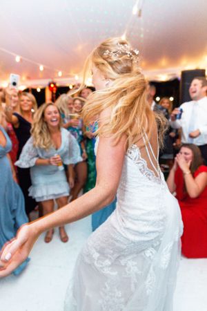 A bride dancing joyfully at her wedding reception under a white tent with guests cheering around her.