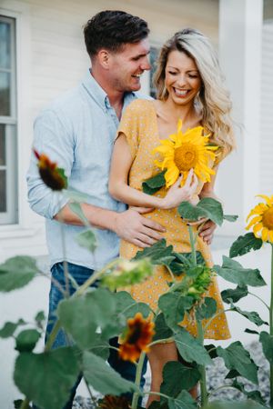 Smiling couple embracing among sunflowers, woman in yellow dress holding bright sunflower bouquet.